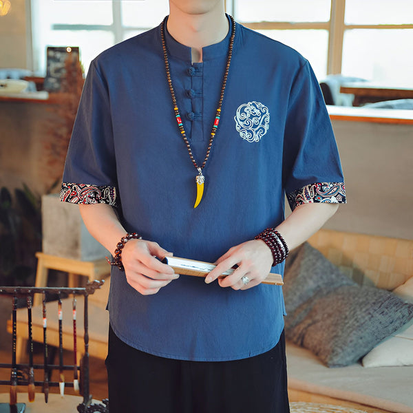Homme portant une chemise bleue à manches courtes avec motif brodé, un collier en perles et tenant un éventail. Détails de motifs paisley aux manches.