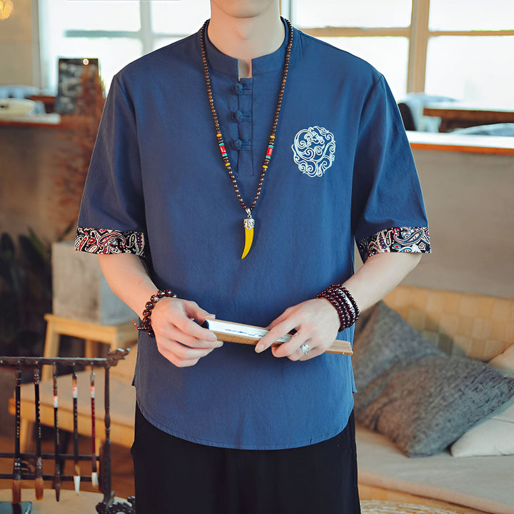 Homme portant une chemise bleue à manches courtes avec motif brodé, un collier en perles et tenant un éventail. Détails de motifs paisley aux manches.