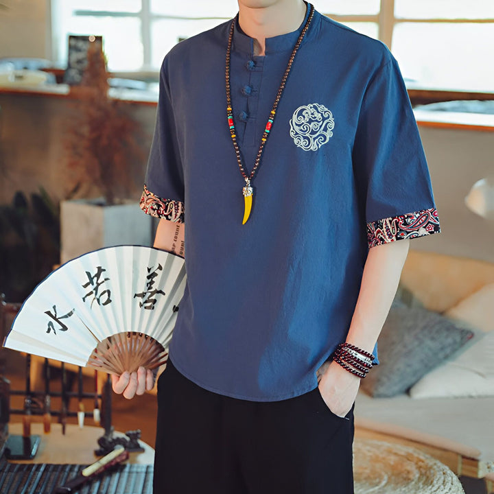Homme en chemise bleue de style chinois avec un éventail. La chemise a des broderies et des bordures à motifs. L'homme porte un collier avec un pendentif jaune.