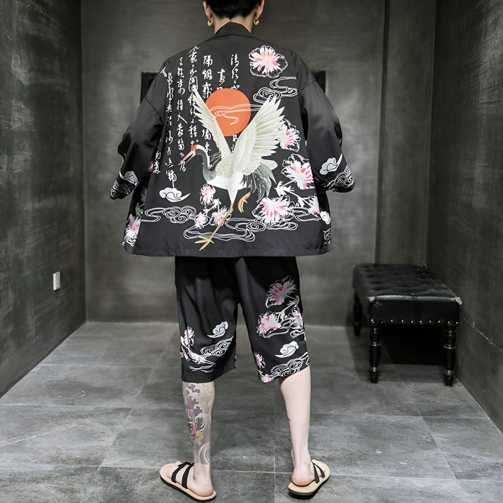 Homme portant un kimono noir et un short assorti, avec un motif de grue blanche, de fleurs roses et d'un soleil rouge. Vêtements de style japonais, vente de kimonos en ligne.