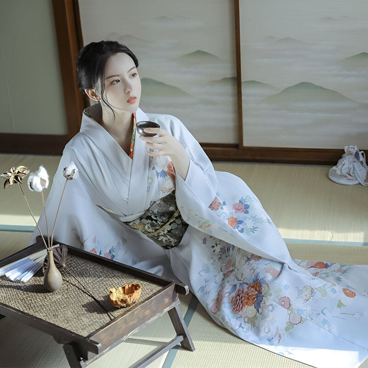 Femme portant un kimono blanc avec motifs floraux, assise, tenant une tasse de thé. Vente de kimonos traditionnels japonais.