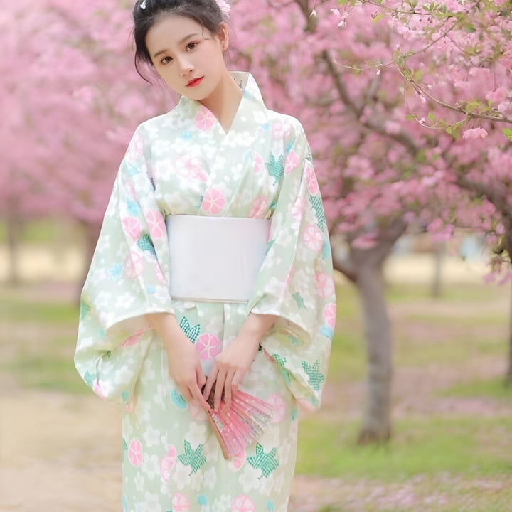 Femme portant un kimono traditionnel vert clair à motifs floraux sous un arbre en fleurs. Kimonos authentiques à vendre.