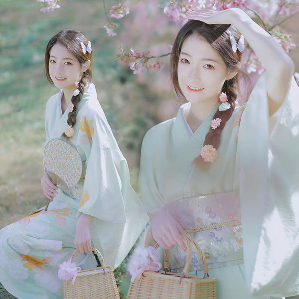 Femme souriante portant un kimono vert clair avec des fleurs, assise devant des cerisiers en fleurs. Vente de kimonos traditionnels et modernes.