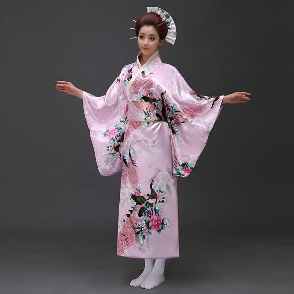 Femme en kimono rose avec motifs de paons et de fleurs. Coiffure traditionnelle japonaise avec un éventail. Elle pose avec les bras tendus.