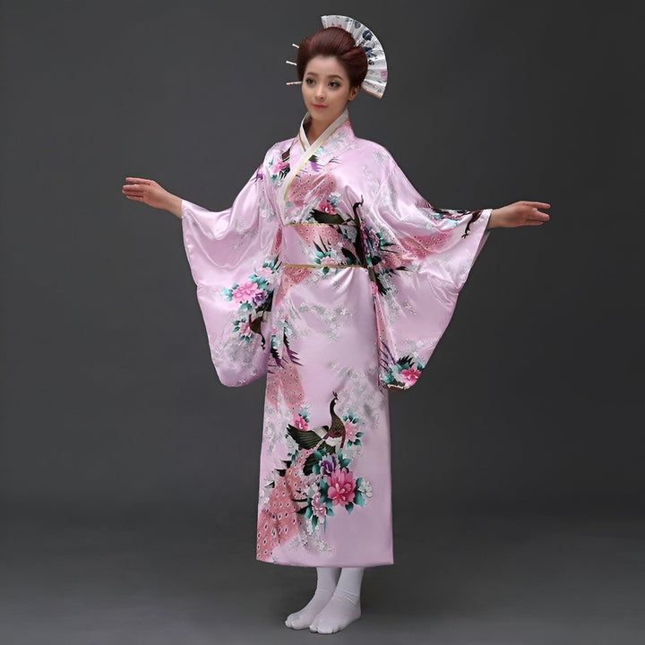 Femme en kimono rose avec motifs de paons et de fleurs. Coiffure traditionnelle japonaise avec un éventail. Elle pose avec les bras tendus.