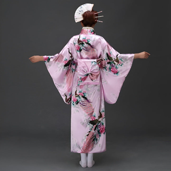 Vue arrière d'une personne en kimono rose à motifs de paons et fleurs, tenant un éventail blanc.