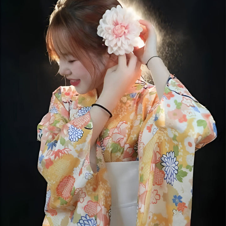 Jeune femme souriante portant un kimono traditionnel fleuri jaune et orange, avec une fleur rose dans les cheveux. Vêtements japonais à vendre.