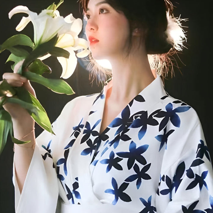 Femme portant un kimono blanc à motifs floraux bleus, tenant un lys blanc. Élégance et tradition japonaises. Découvrez notre collection de kimonos.
