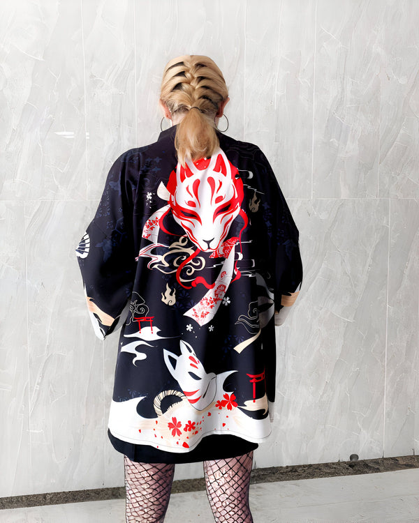 Femme vue de dos portant un kimono noir imprimé de motifs japonais, notamment des masques de renard rouges et blancs. Kimono unique disponible dans notre boutique en ligne.