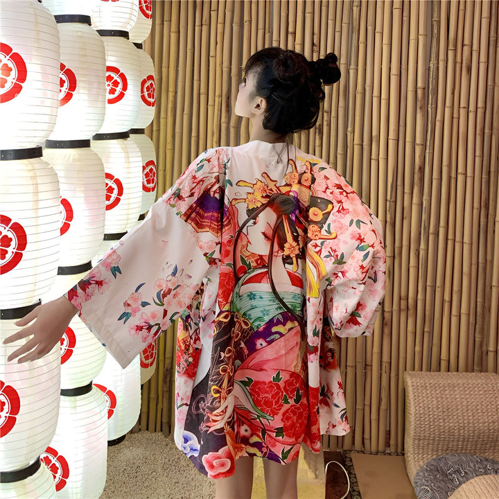 Femme portant un kimono blanc avec imprimé d'une geisha et de fleurs de cerisier, devant un mur de lanternes japonaises. Vente de kimonos en ligne.