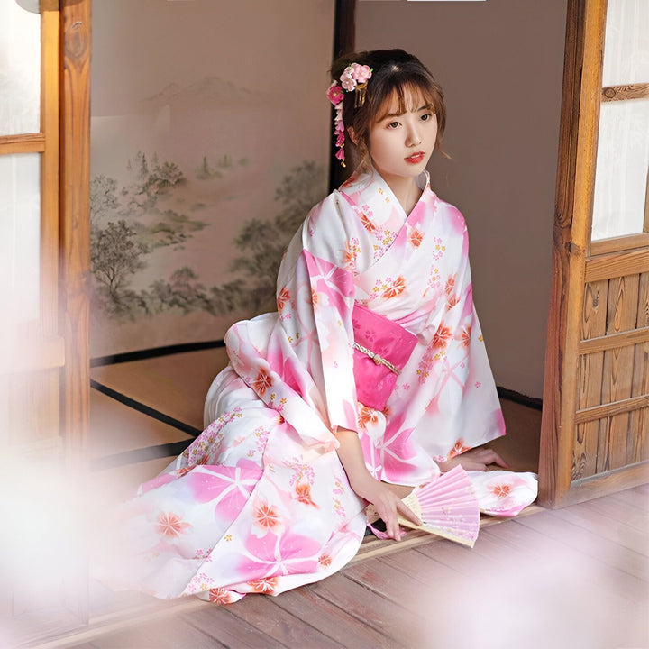 Femme asiatique en kimono blanc et rose à motifs floraux, assise sur un tatami. Accessoire floral dans les cheveux. Kimono traditionnel japonais à vendre.