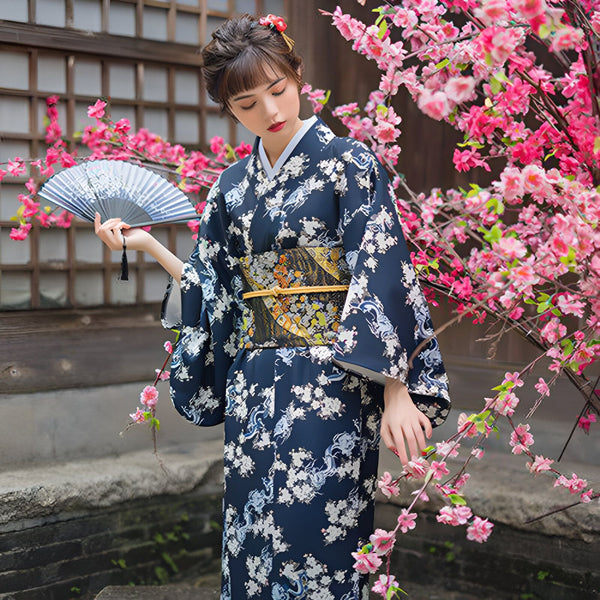 Femme en kimono bleu marine fleuri, tenant un éventail. Arbre en fleurs roses en arrière-plan. Vêtements japonais traditionnels.