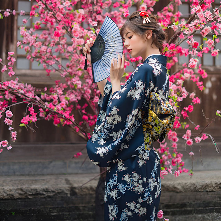 Femme élégante en kimono bleu marine à motifs floraux, tenant un éventail devant un cerisier en fleurs. Vente de kimonos authentiques.