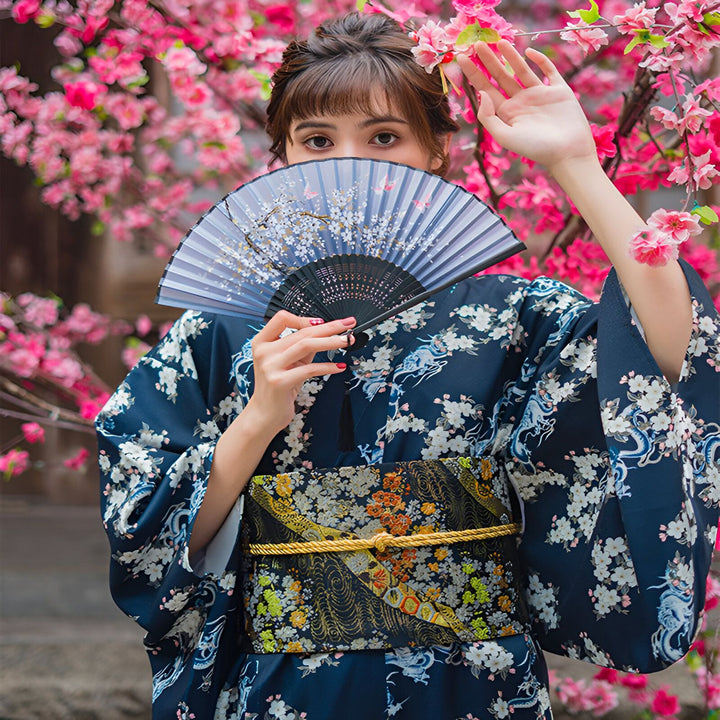 Jeune femme en kimono bleu marine à motifs floraux, tenant un éventail orné de fleurs de cerisier. Arrière-plan de fleurs roses. Vente de kimonos.