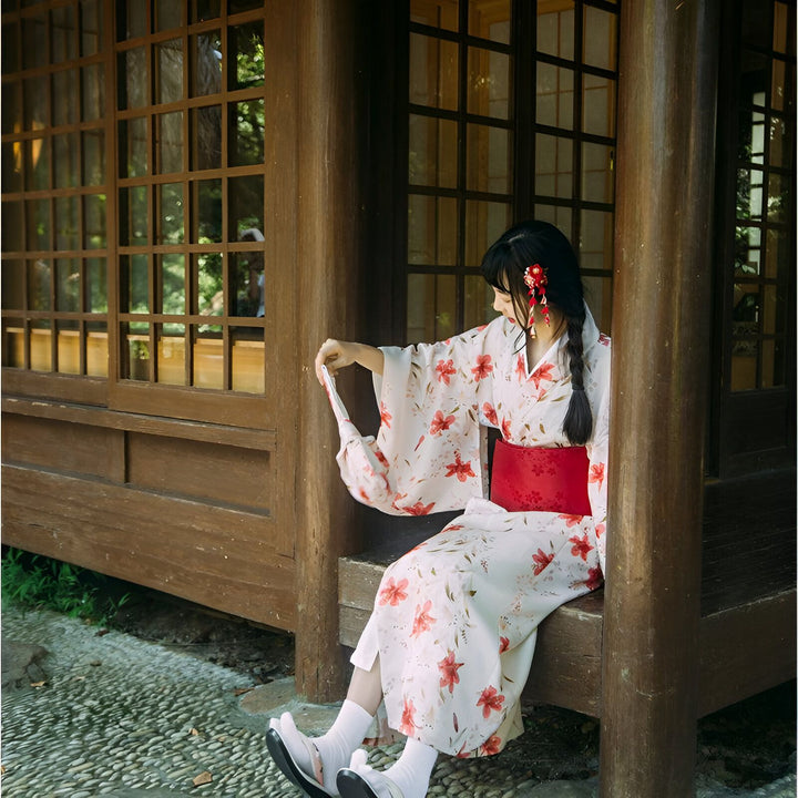 Femme en kimono fleuri, assis devant une maison en bois traditionnelle japonaise. Kimonos de qualité à découvrir.