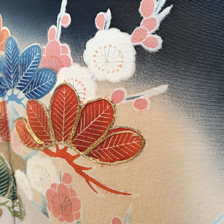 Détail d'un kimono de luxe avec motifs floraux japonais complexes : feuilles brodées rouge et or, fleurs de prunier blanches et roses sur fond dégradé bleu marine et beige. Parfait pour les amateurs de kimonos et de culture japonaise.