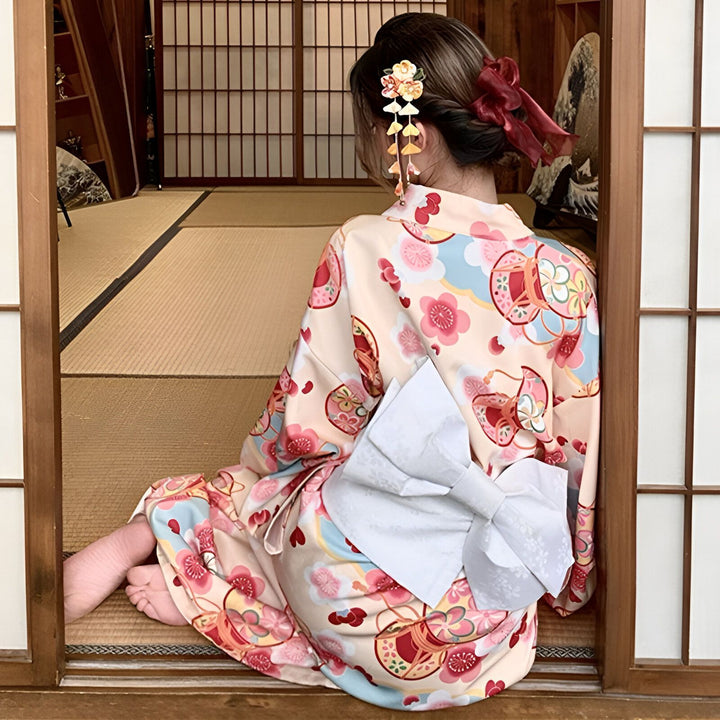 Femme portant un kimono traditionnel à motifs floraux, vue de dos. Coiffure ornée d'une fleur et d'un ruban rouge. Kimonos disponibles à l'achat.