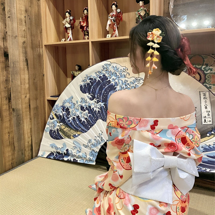 Femme portant un kimono traditionnel japonais à motifs floraux, avec une épingle à cheveux décorative. En arrière-plan, une estampe de vague et une étagère avec des poupées japonaises. Vente de kimonos authentiques.