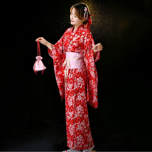 Une femme souriante en kimono rouge à motifs de fleurs blanches, ceinturée de rose, tenant une bourse. Coiffure ornée de fleurs et ruban rouges. Fond noir.