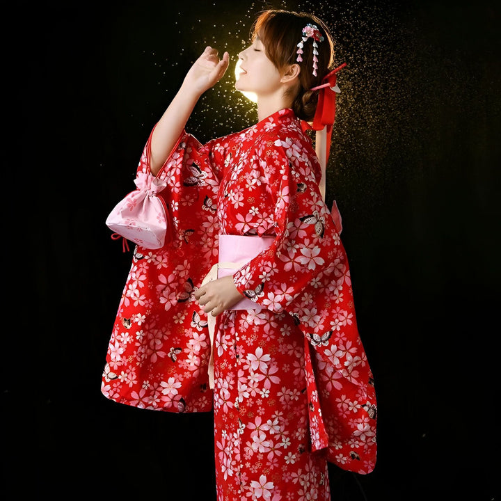 Jeune femme en kimono rouge à motifs floraux, accessoire de cheveux fleuri et ruban rouge, posant avec grâce sous une lumière dorée. Beauté asiatique, tradition japonaise, élégance et raffinement.