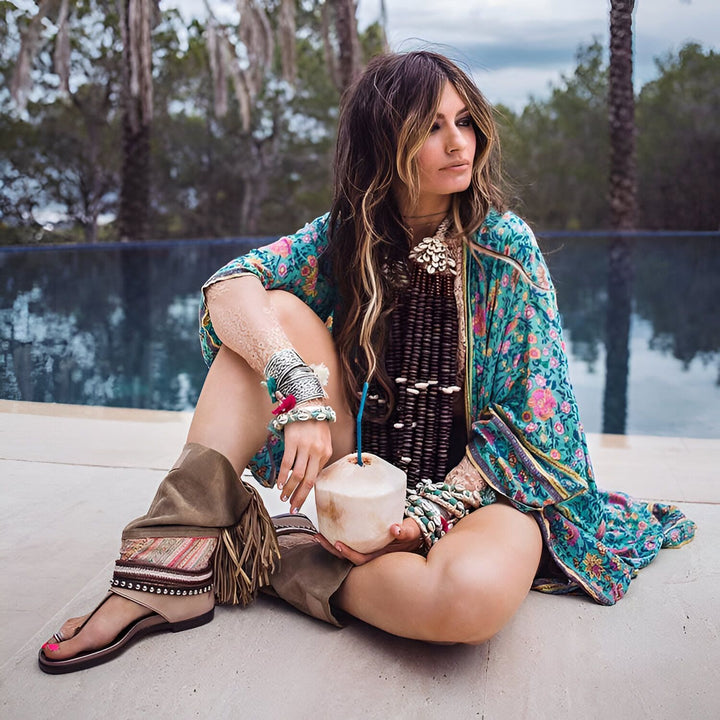 Femme portant un kimono fleuri turquoise et des accessoires bohèmes. Elle tient une noix de coco près d'une piscine. Collection de kimonos pour un style unique. Découvrez nos kimonos.