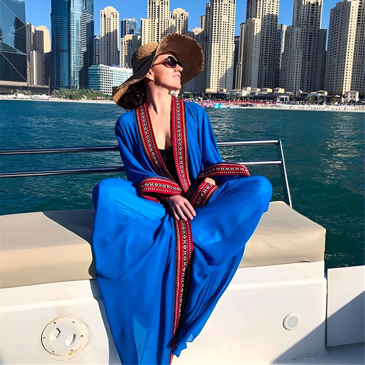 Femme élégante sur un bateau, portant un kimono bleu vif avec bordure rouge. Chapeau de paille et lunettes de soleil. Paysage urbain en arrière-plan. Kimonos de luxe disponibles.