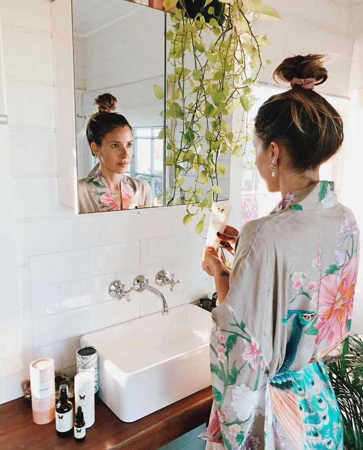 Femme en kimono fleuri dans salle de bain. Miroir reflétant le visage, lavabo blanc, plante verte suspendue. Produits de beauté visibles, ambiance cocooning. Kimonos fleuris pour moments de détente.