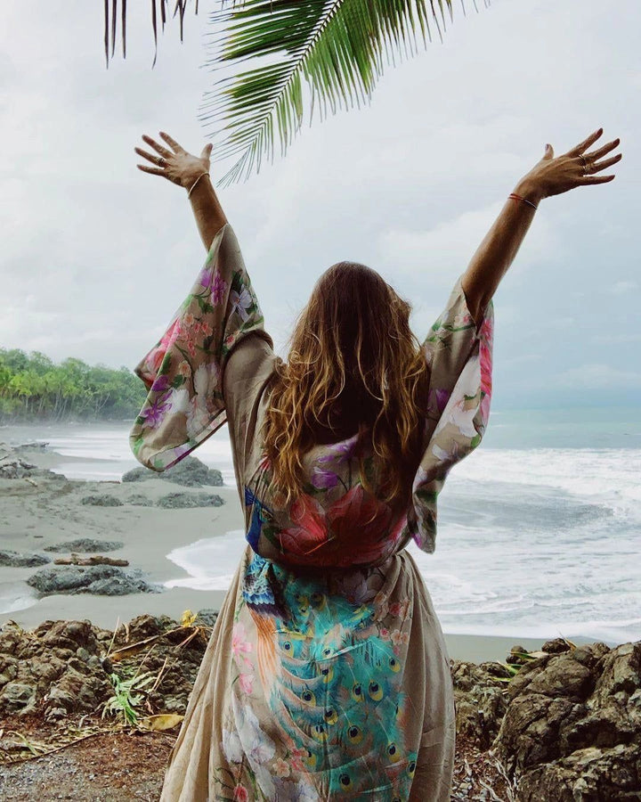 Femme portant un kimono à motifs floraux et paon, bras levés face à l'océan. Vêtement de style bohème, disponible à l'achat.