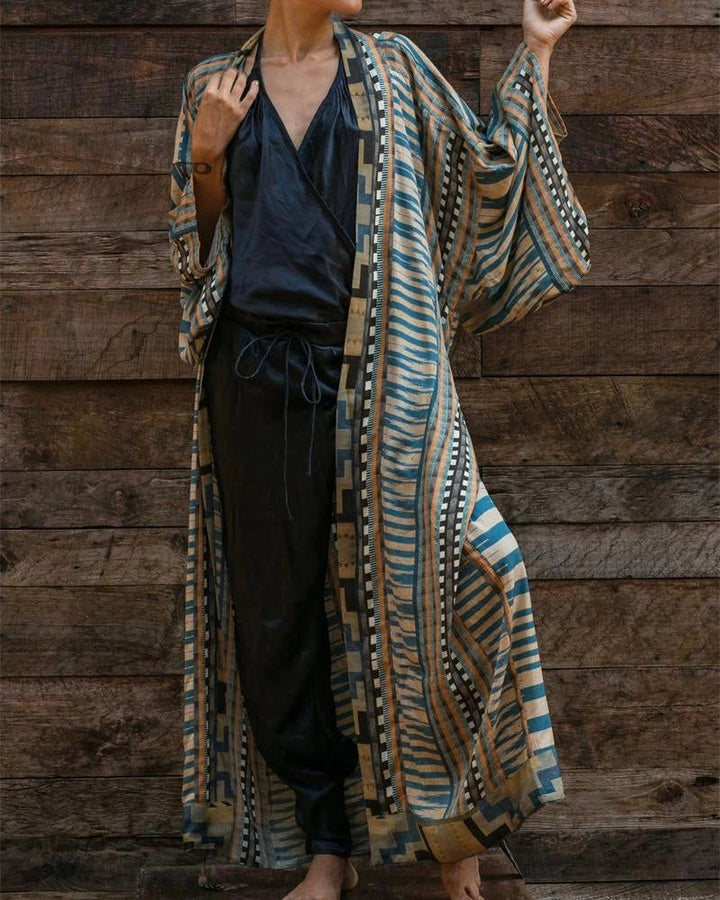 Femme portant un kimono long à motifs bleus et beiges devant un mur en bois. Vêtement fluide et élégant, parfait pour la détente ou une occasion spéciale. Découvrez notre collection de kimonos uniques.