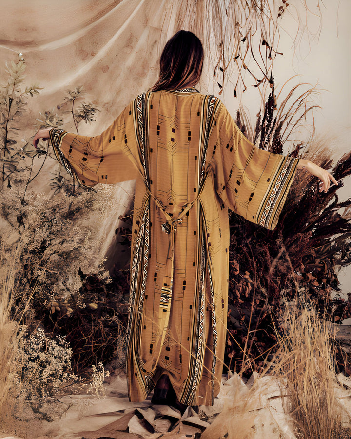 Femme de dos portant un kimono long ocre avec motifs géométriques noirs et blancs, ceinture nouée. Notre collection de kimonos offre des pièces uniques pour un style bohème et élégant. Découvrez notre sélection en ligne.