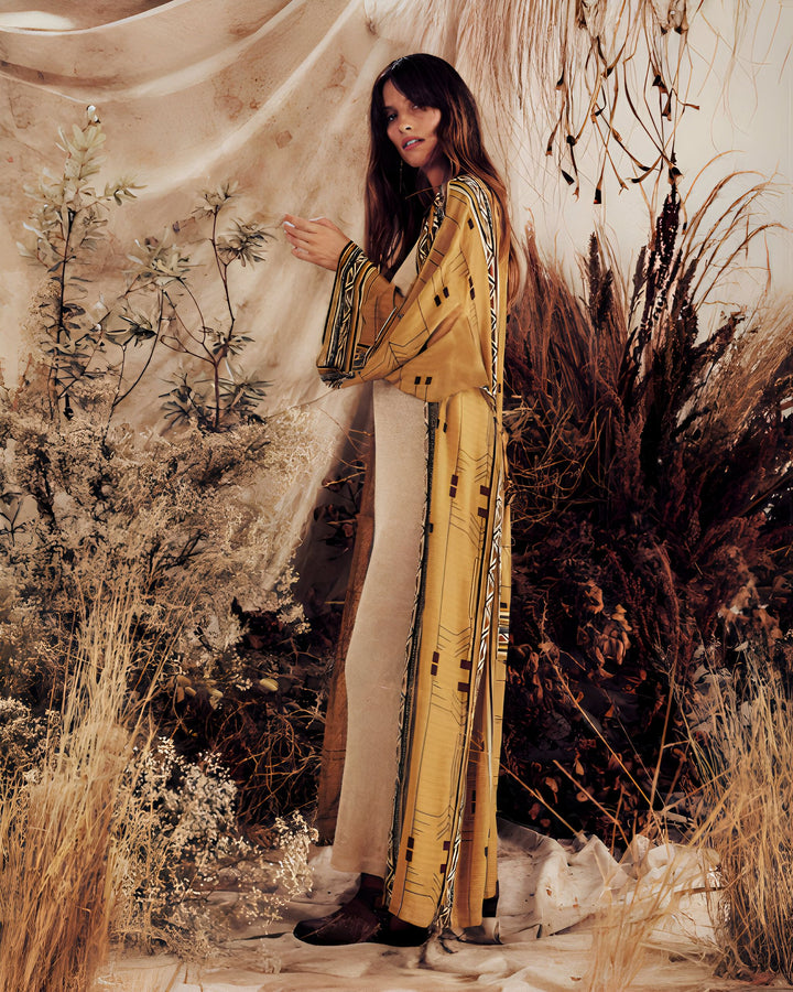 Femme portant un kimono long jaune moutarde avec motifs géométriques. Style bohème, pose naturelle devant un décor végétal texturé. Vêtements kimono tendance.