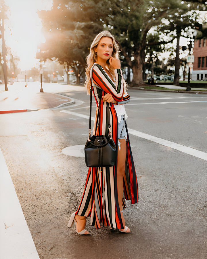Femme blonde portant un kimono long à rayures colorées, short en jean, sac en cuir noir. Vente de kimonos originaux en ligne. Style estival tendance et décontracté.