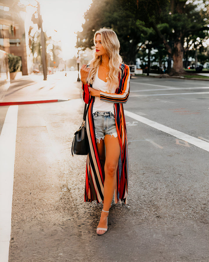 Femme blonde portant un kimono long à rayures colorées, un short en jean et un débardeur blanc. Sac à main noir et talons hauts. Look tendance et décontracté. Kimonos disponibles à la vente.