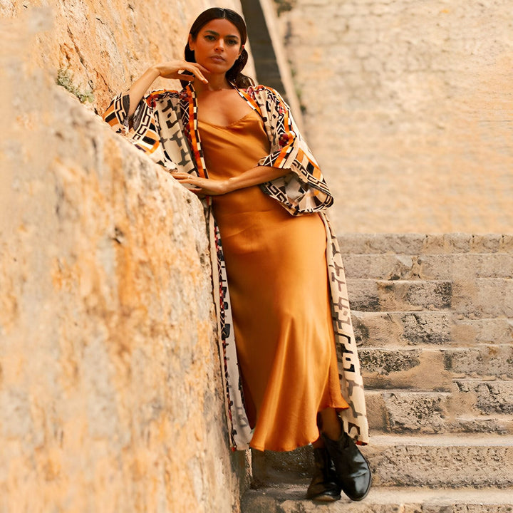 Femme portant un kimono à motifs géométriques sur une robe dorée, adossée à un mur en pierre. Collection de kimonos pour femmes.