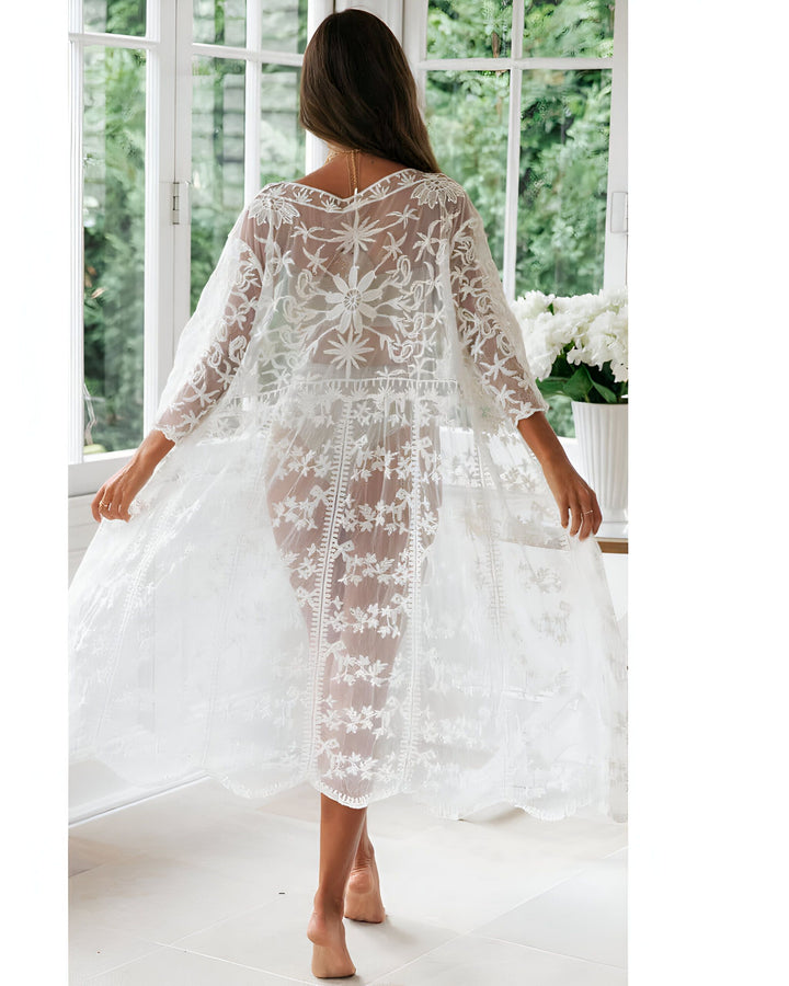 Femme portant un kimono long en dentelle blanche devant une fenêtre. Kimono transparent, motif floral délicat. Découvrez notre collection de kimonos élégants.