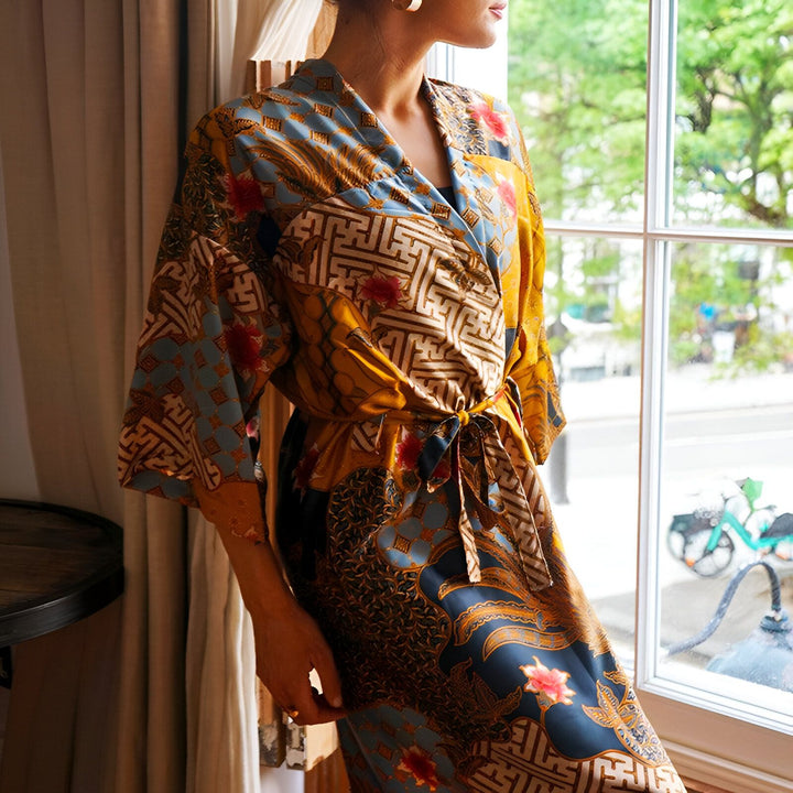 Femme portant un kimono coloré à motifs devant une fenêtre. Élégance et confort réunis. Découvrez notre collection de kimonos uniques.
