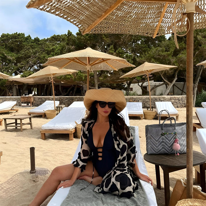 Femme à la plage portant un kimono, un maillot de bain noir et un chapeau de paille. Parasols et chaises longues en arrière-plan. Vente de kimonos en ligne.
