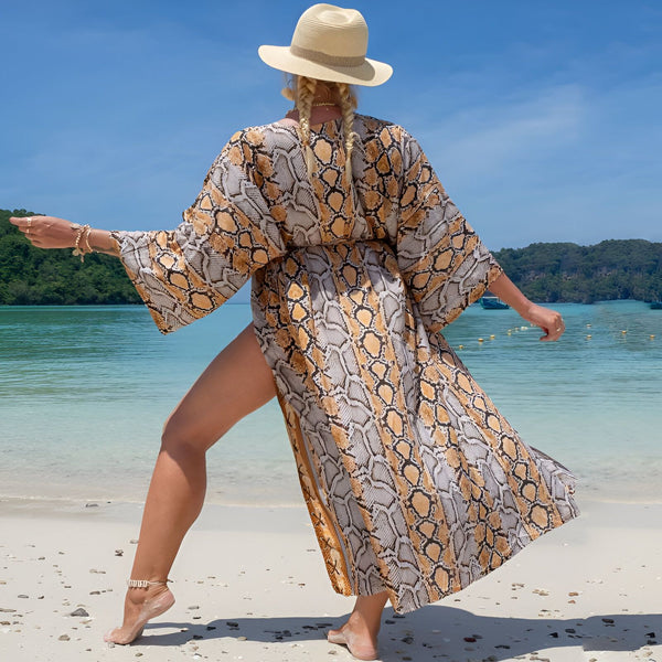 Femme portant un kimono à motif serpent sur une plage ensoleillée. Kimono long et fluide avec fente latérale. Chapeau de paille et bracelet. Mode plage, style bohème.