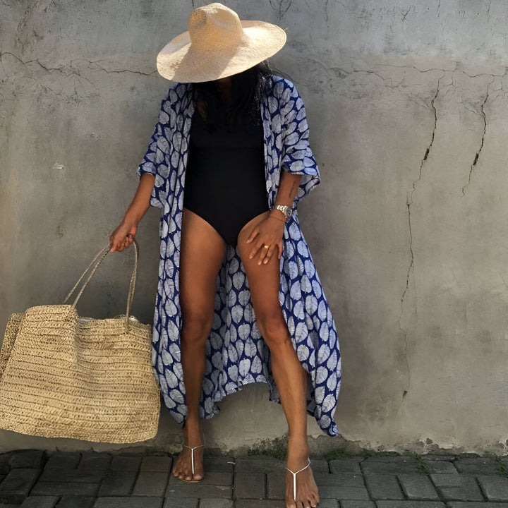 Femme portant un kimono imprimé bleu et blanc par-dessus un maillot de bain noir, avec un chapeau de paille et un sac de plage en paille. Kimonos tendance pour la plage.