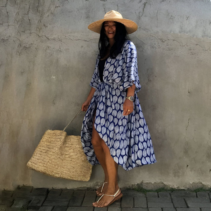 Femme souriante portant un kimono bleu à motifs de feuilles, chapeau de paille et sac en fibres naturelles. Vente de kimonos pour femmes, styles variés.