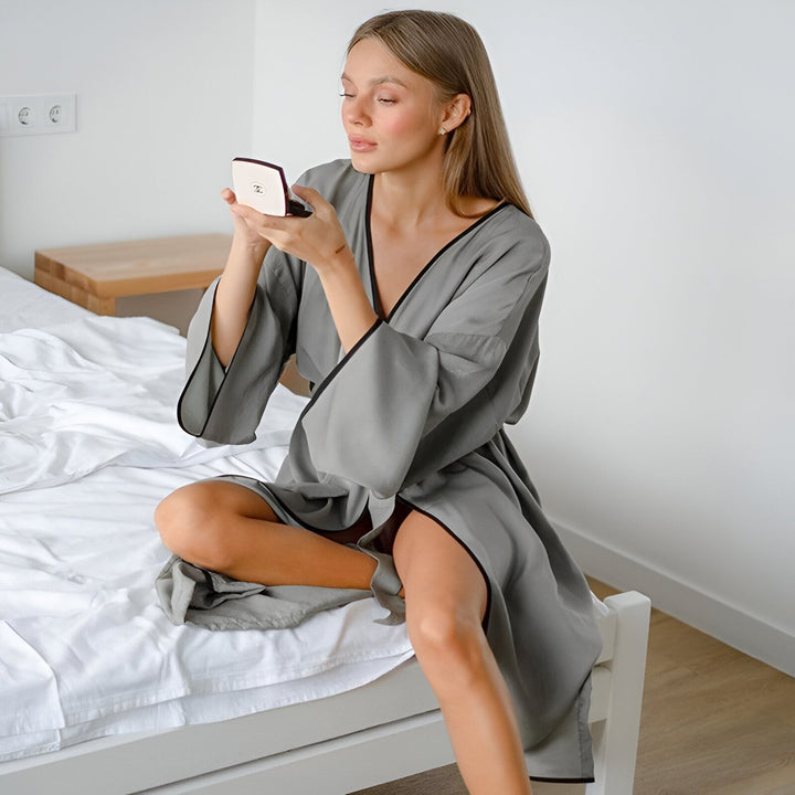Femme assise sur un lit, portant un kimono gris avec bordure noire. Elle se maquille avec un miroir à main. Vente de kimonos en ligne.