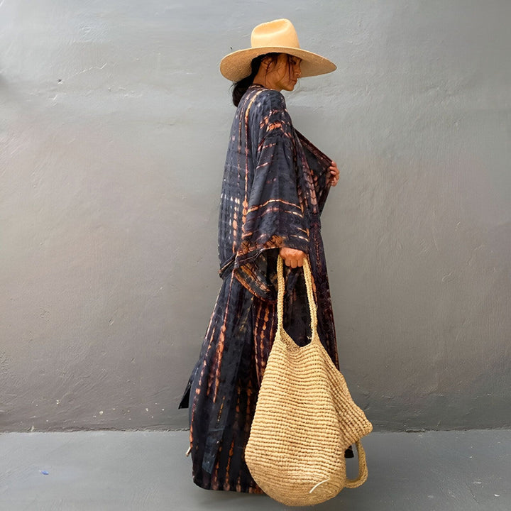 Femme portant un kimono long à motifs tie-dye, chapeau de paille et sac en fibres naturelles. Kimonos de créateurs, look bohème chic.