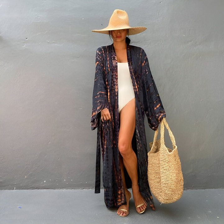 Femme portant un kimono long tie-dye noir et orange par-dessus un maillot de bain blanc. Elle porte un chapeau de paille et un sac en osier. Kimonos tendance en vente en ligne.