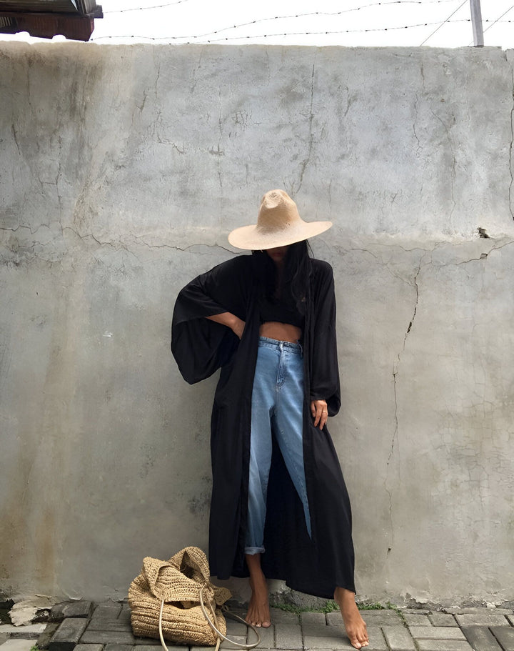 Femme portant un kimono noir long, jeans bleus et chapeau de paille devant un mur en béton. Sac en osier à ses pieds. Découvrez nos kimonos élégants.