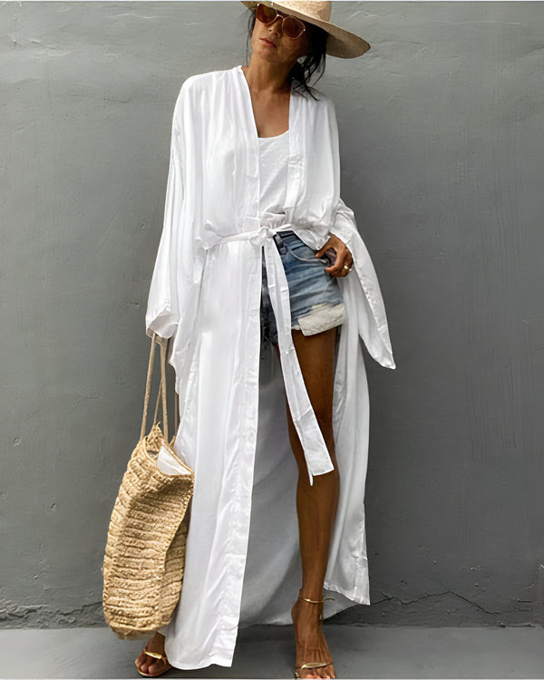 Femme portant un kimono blanc long et fluide. Style décontracté avec short en jean et chapeau de paille. Sac en paille et lunettes de soleil. Kimonos élégants et confortables disponibles dans notre boutique.