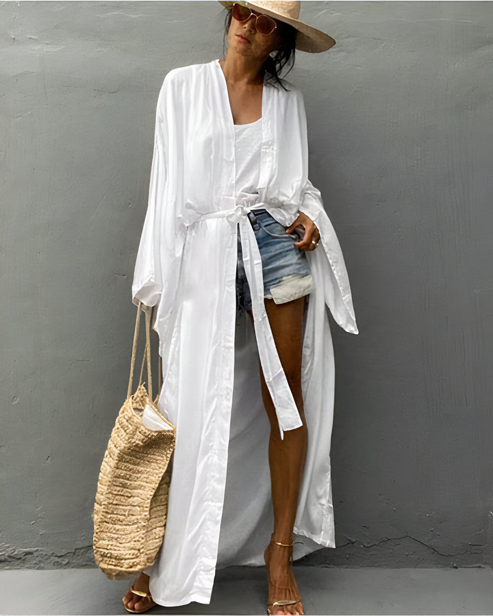 Femme portant un kimono blanc long et fluide. Style décontracté avec short en jean et chapeau de paille. Sac en paille et lunettes de soleil. Kimonos élégants et confortables disponibles dans notre boutique.