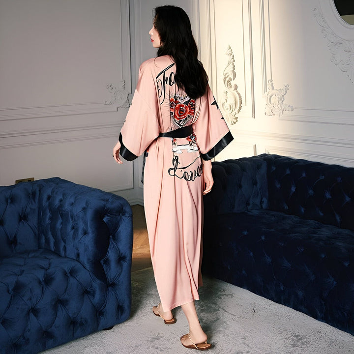 Femme portant un kimono rose avec motifs imprimés, manches amples bordées de noir. Ceinture noire soulignant la taille. Vêtements de nuit élégants, robes de chambre japonaises.