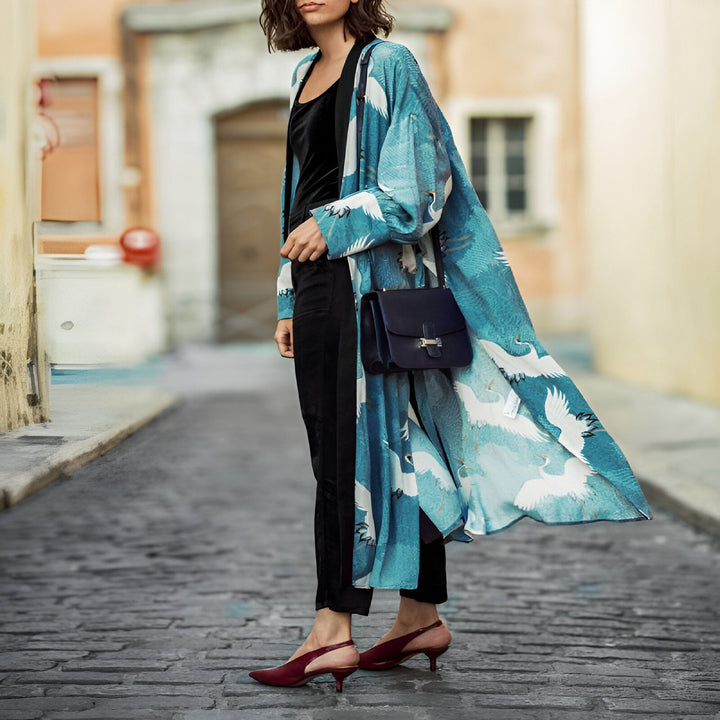 Femme portant un kimono bleu à motifs de grues blanches, pantalon et haut noirs, sac bleu et chaussures bordeaux. Mode kimono tendance.