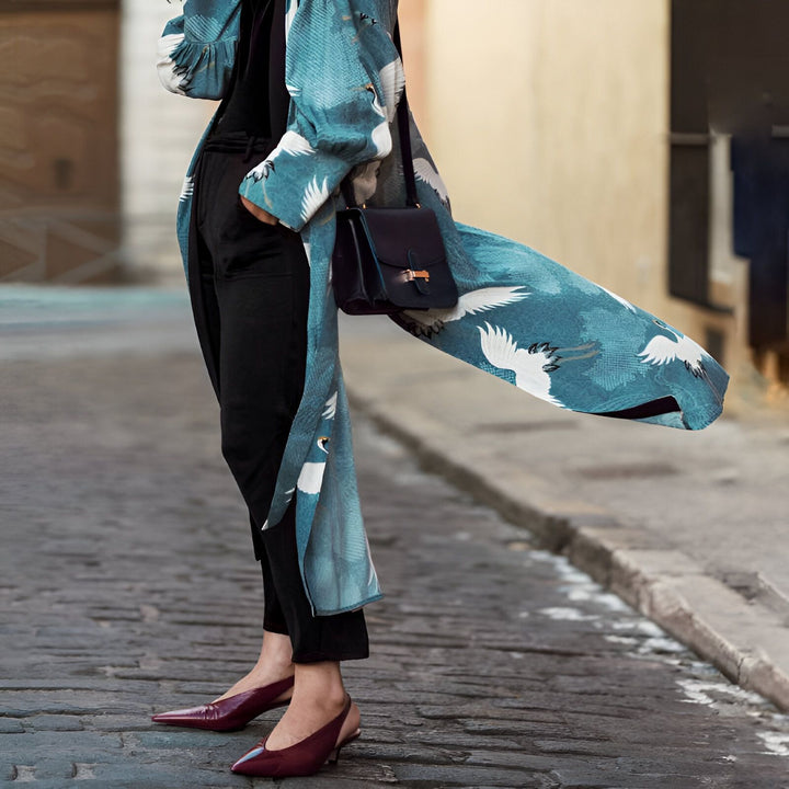Femme portant un kimono bleu à motifs de grues, avec pantalon noir et sac à main. Élégance décontractée. Vêtements de style japonais. Boutique de kimonos en ligne.
