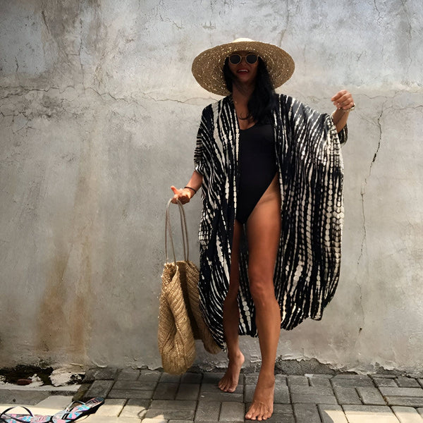 Femme portant un kimono noir et blanc à motifs, un maillot de bain noir, un chapeau de paille et des lunettes de soleil. Elle tient un sac de paille. Vente de kimonos.